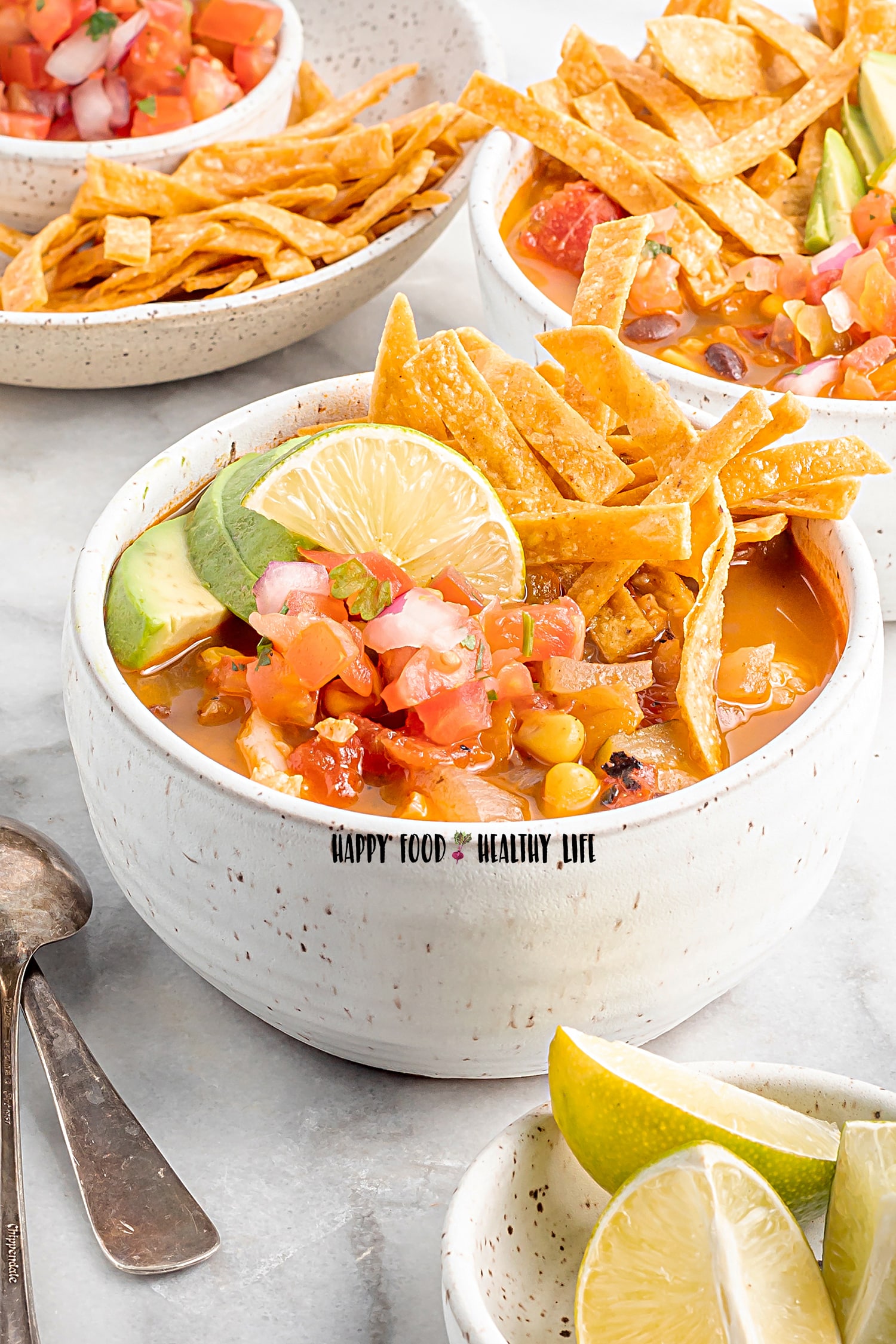 Photo of two white bowls filled with Vegan Tortilla Soup and topped with fresh lime wedges and tortilla strips. There is a bowl of lime wedges in the foreground, as well as a silver spoon, and and a bowl of tortilla strips and salsa in the background.