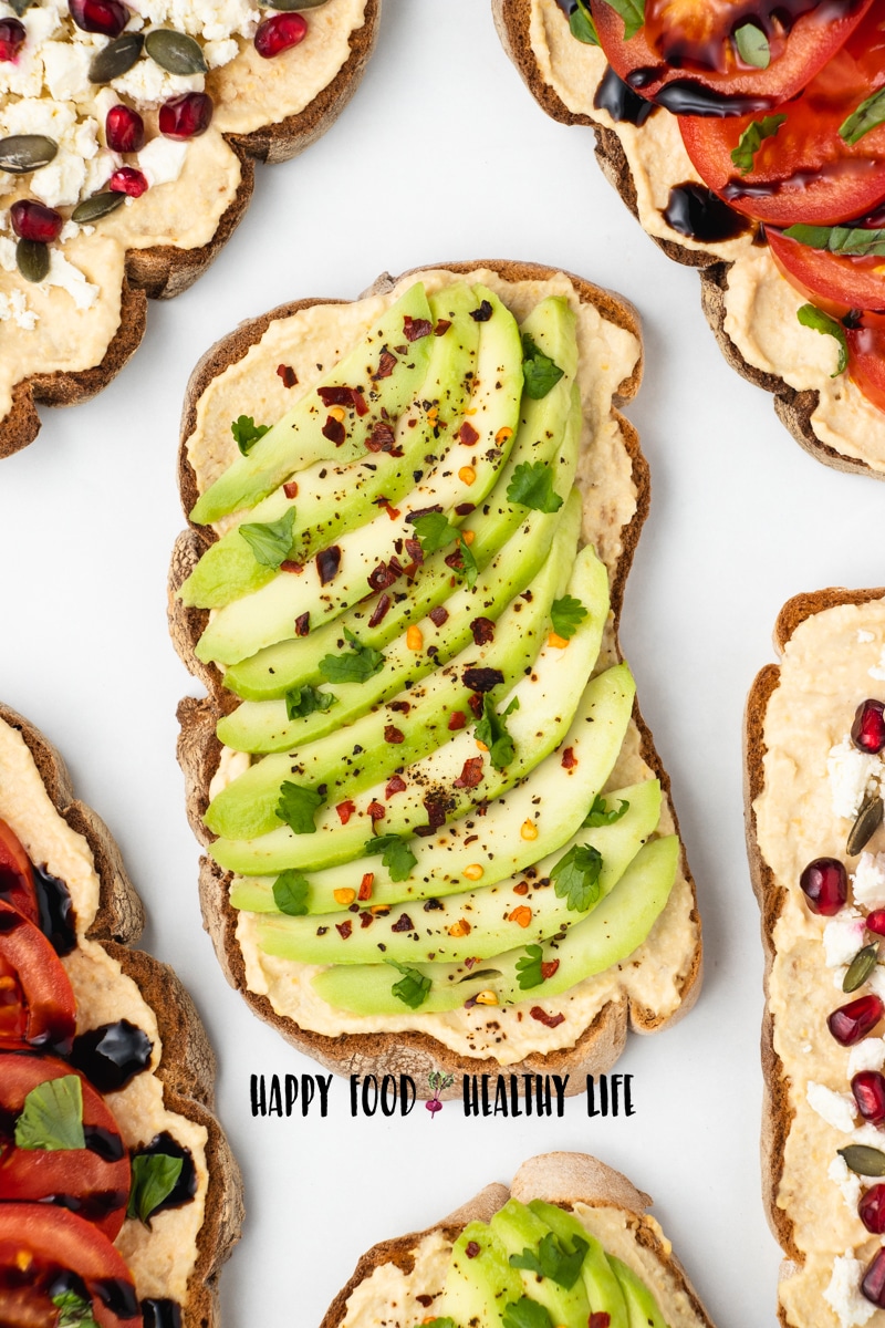 Top view photo of multiple slices of bread with hummus on each slice. The hummus toast is topped with different toppings, including avocado, salt, pepper, cilantro, pomegrante seeds, tomatoes, basil leaves, pumpkin seeds, and balsamic reduction.
