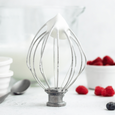 a metal whisk from a stand mixer, standing on a counter showing aquafaba whipped cream with stiff peaks. In the background is a bowl of fresh berries.