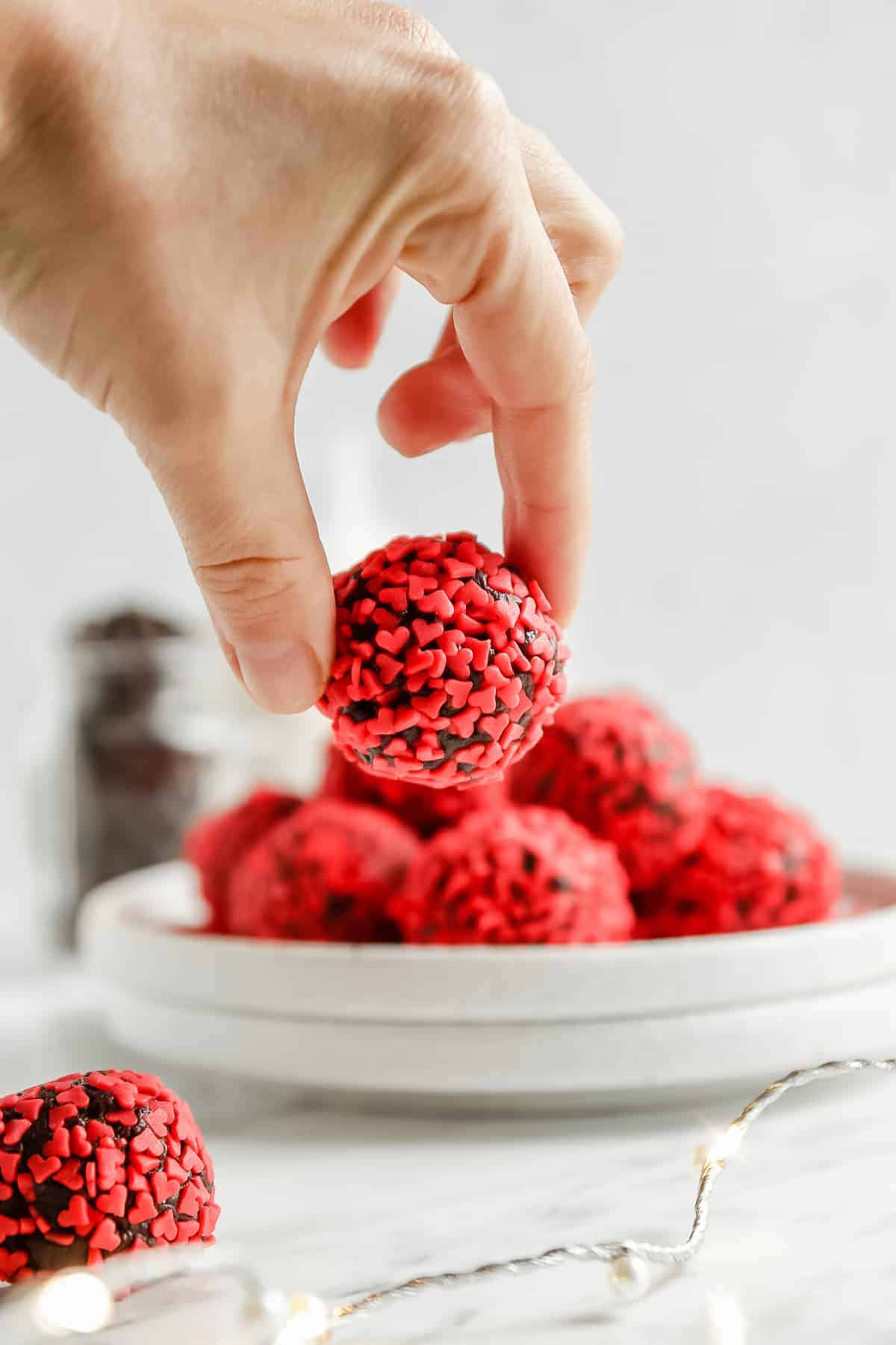 a close-up of a valentine's truffle being held by two fingers. in the background, there is a plate of many truffles.