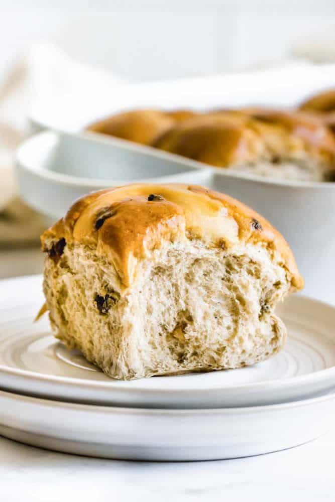 Single Vegan Hot Cross Bun on white plate with dish of buns in the background.