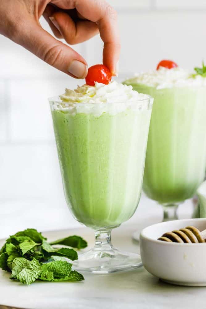 A hand placing a cherry on top of a glass of dairy-free shamrock shake.