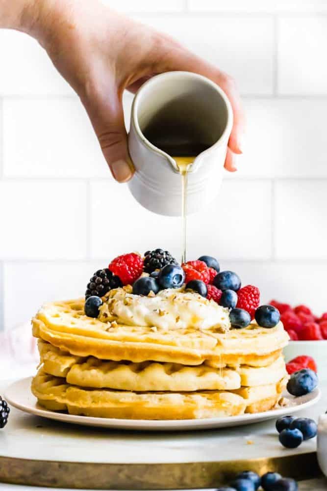 Hand pouring syrup over the best vegan waffles topped with vegan cream and fresh berries.