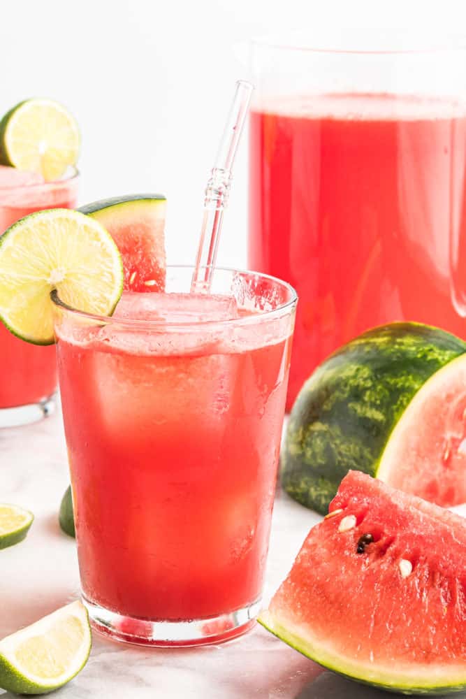 Glass of watermelon agua fresca with straw and lime slice garnish.  Watermelon slices in background.