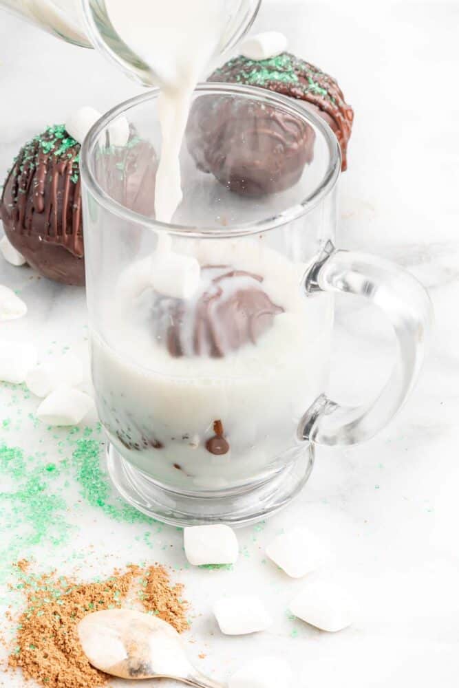 pouring hot milk over a vegan hot chocolate bomb in a glass mug. 
