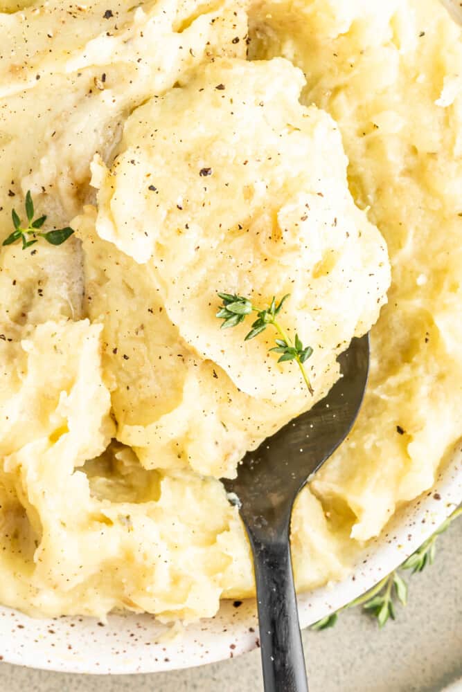 a big spoonful of vegan mashed potatoes in a ceramic dish. potatoes are topped with fresh herbs and pepper.