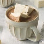 an off white ceramic mug of hot cocoa topped with two square vegan marshmallows.