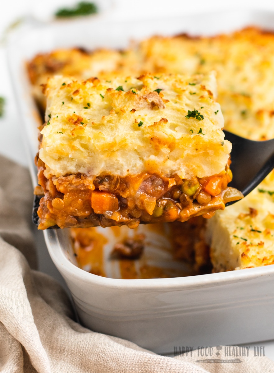 a white casserole dish filled with lentil shepherd's pie. A spatula is holding up a corner pie to show the layer of lentils and veggies under a thick layer of mashed potatoes. Photo Credit: Cindy Gordon of HappyFoodHealthyLife.com
