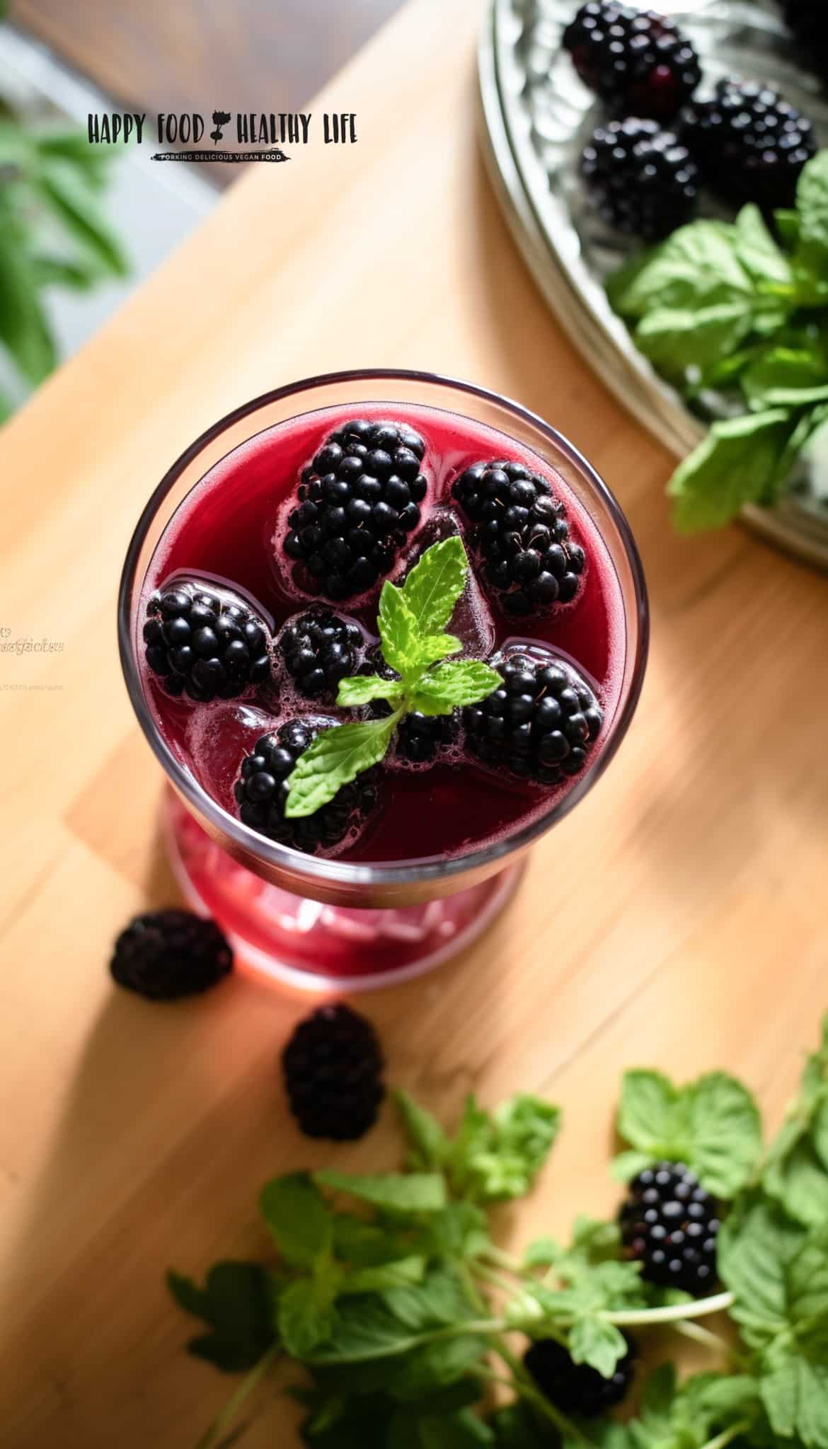 overhead top to bottom shot of a clear glass filled with thin purple liquid with blackberries and mint in the liquid and blackberries and mint beside the glass