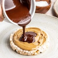 Photo of a rice cake, topped with peanut butter, with creamy dark chocolate being poured on top.