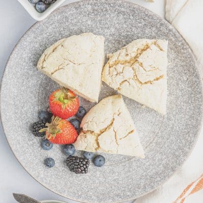 Top view photo of three vegan scones, on a textured gray plate, with fresh fruit like strawberries, blueberries, and blackberries.