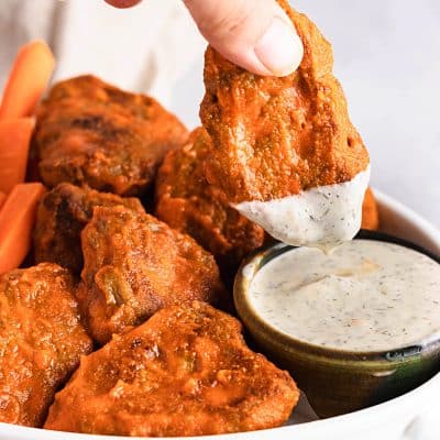 Closeup photo of a white bowl filled with vegan buffalo wings. There is a hand holding one of the wings, and dipping it into a small black bowl filled with vegan ranch dressing. There are also carrot sticks in the back of the bowl.
