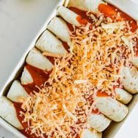 Photo of enchiladas in a white casserole dish, topped with Vegan Mexican cheese blend, and ready to bake. There are lime wedges next to the dish.
