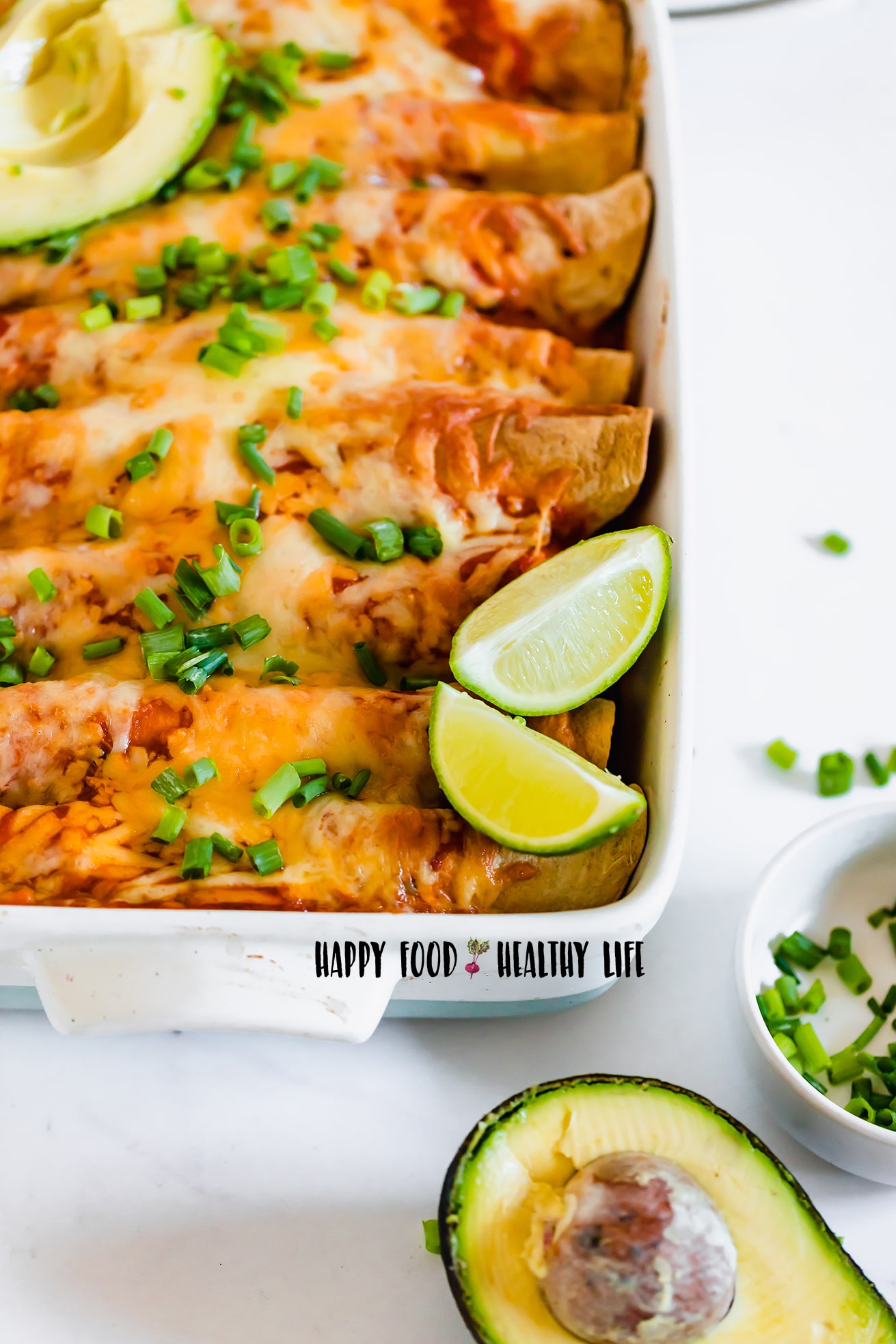 Photo of a white casserole dish, filled with Vegan Enchiladas, ready to enjoy. the entire dish is topped with fresh avocado, lime wedges, and chopped spring onions.
