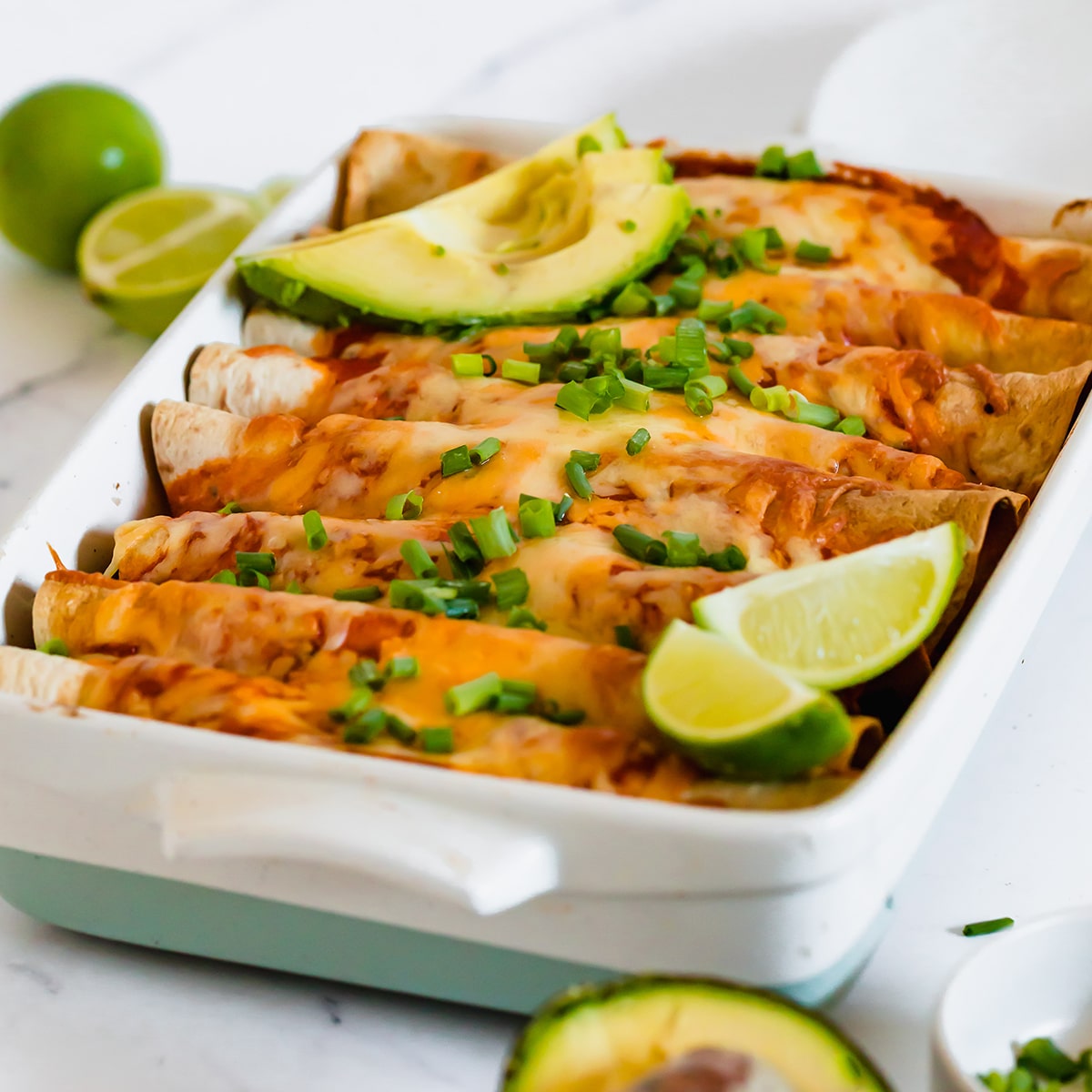 Photo of a white casserole dish filled with vegan enchiladas, and topped with fresh lime wedges, avocado slices, and chopped spring onions.