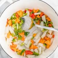 Top view photo of a white stock pot, with chopped veggies in the pot. There is a large silver spoon in the pot, mixing the veggies. The veggies include onions, bell peppers, and jalapeno peppers.