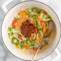 Top view photo of a white stock pot, with chopped veggies and spices in the pot. There is a large silver spoon in the pot, mixing the veggies. The veggies include onions, bell peppers, and jalapeno peppers. And the spices that were added in are chili powder and garlic.