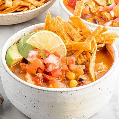 Closeup photo of two white bowls filled with Vegan Tortilla Soup and topped with fresh lime wedges and tortilla strips. There is a bowl of tortilla strips and salsa in the background.