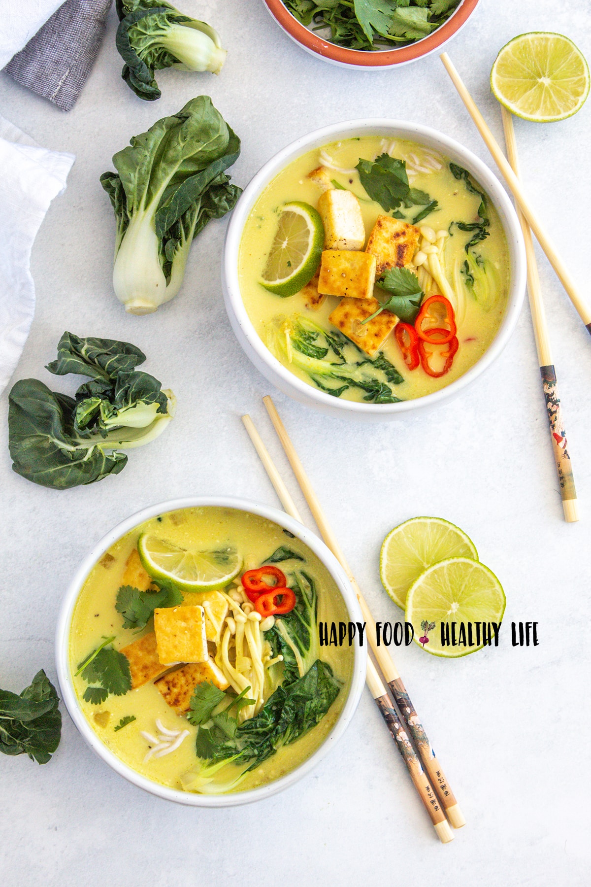 Top view photo of two white bowls filled with vegan ramen soup with tofu. Surrounding the bowl there are lime slices, chopsticks, and bok choy. 