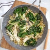 Top view photo of bok choy and mushroom in the saucepan with the onions and garlic. The pan is sitting on a wooden cutting board and there is a white kitchen towel next to the pan.