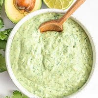 Top view photo of a white bowl filled with avocado cream sauce. There is a wooden spoon in the bowl and the bowl is surrounded by fresh cilantro, avocado halves, and lime halves.