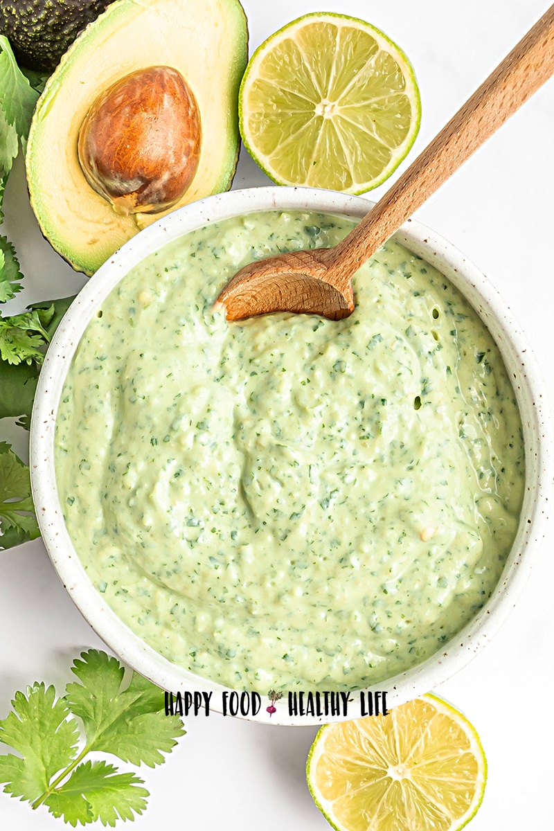 Top view photo of a white bowl filled with avocado cream sauce. There is a wooden spoon in the bowl and the bowl is surrounded by fresh cilantro, avocado halves, and lime halves.