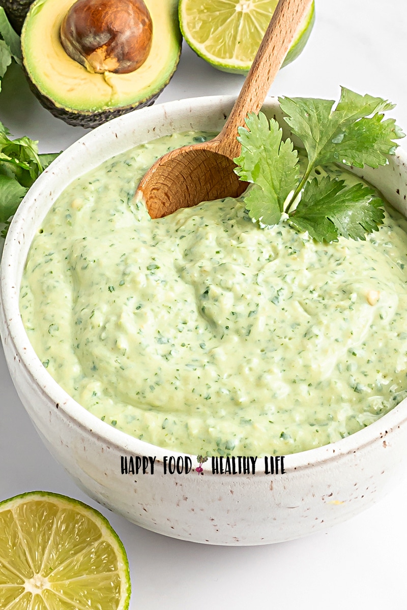 Photo of a white bowl filled with Avocado Cream Sauce. There is fresh cilantro and a wooden spoon in the bowl, and in the foreground there is a lime slice. In the background there is an avocado that is cut in half. 
