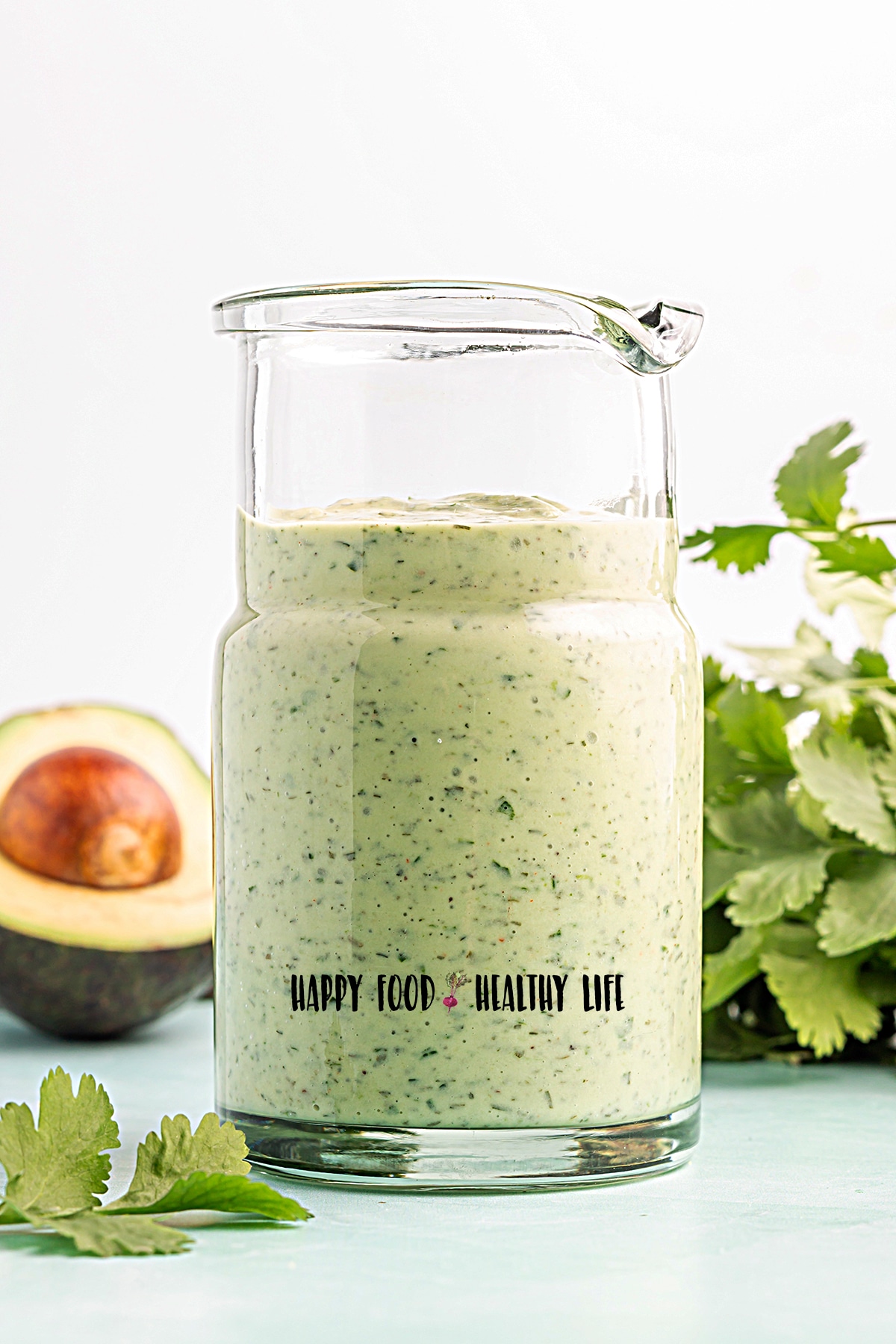 Photo of a glass dressing jar filled with freshly-made avocado lime ranch dressing. In the background there is half an avocado and a bunch of cilantro. 