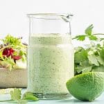 Photo of a glass dressing jar filled with freshly-made avocado lime ranch dressing. In the background there is a white bowl filled with mixed greens, and a bunch of cilantro. In the foreground is a lime sliced in half.