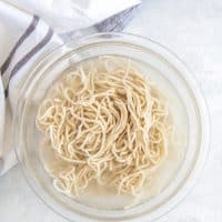 Top view photo of a glass bowl of cooked ramen noodles, that have been set aside until the broth is ready. There is a kitchen towel next to the bowl.