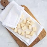 Top view photo of a wooden cutting board, covered with a white kitchen towel and cubed, firm tofu. The excess moisture has been drained as the tofu is pressed into the kitchen towel.