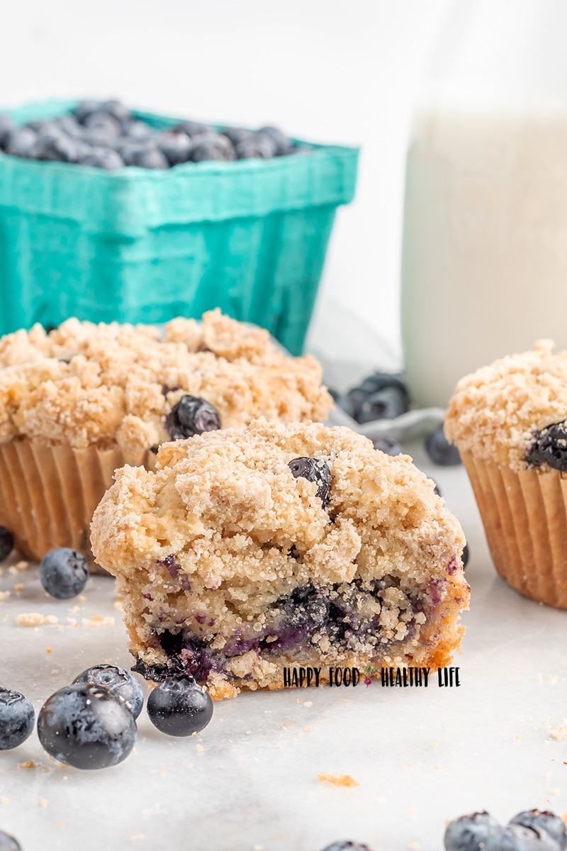 Photo of 3 vegan blueberry muffins. The one in the center has had a bite taken out of it. There are fresh blueberries scattered around the countertop, and a pitcher of milk and teal milk carton with fresh blueberries in the background. 