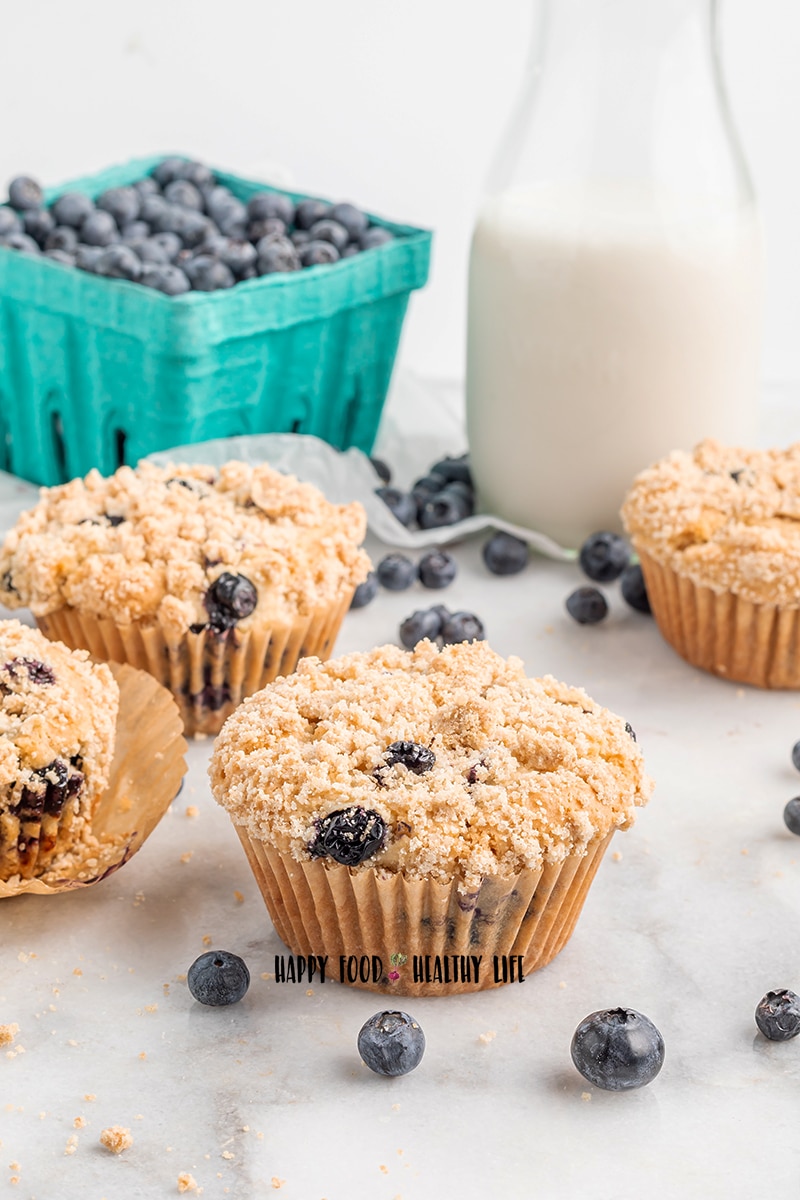 Photo of 4 vegan blueberry muffins. In the foreground are a couple fresh blueberries on the counter. In the background is a glass milk pitcher filled with milk, and a teal blueberry carton filled with fresh blueberries. 