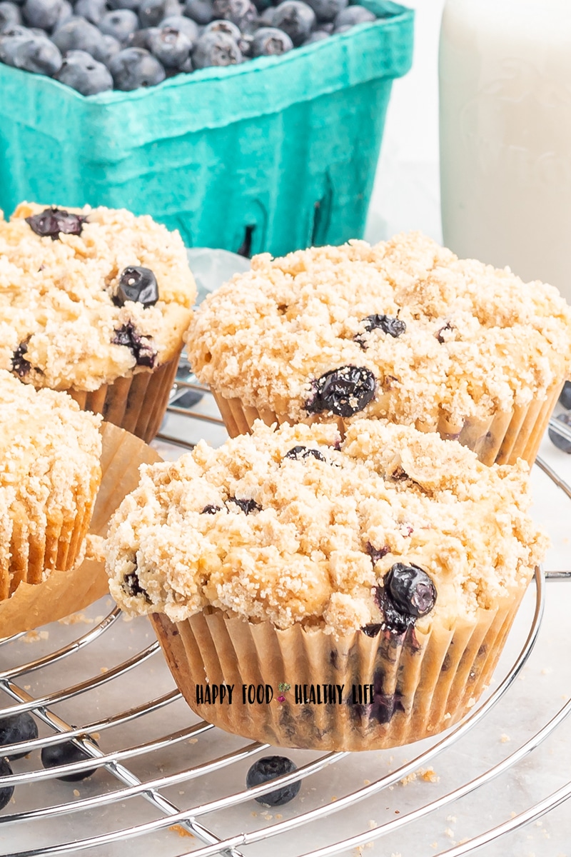 Closeup photo of 4 vegan blueberry muffins on a wire cooling rack. In the background is a glass pitcher of milk and a teal carton of fresh blueberries. 