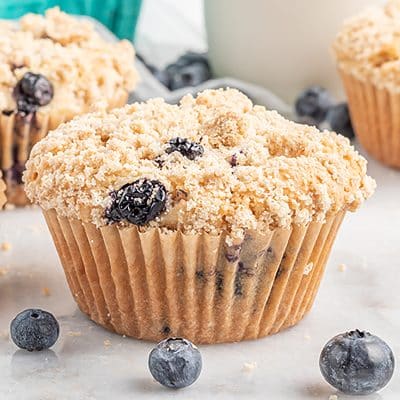 Closeup photo of a vegan blueberry muffin. There are fresh blueberries scattered on the countertop, and muffins and a pitcher of milk in the background.