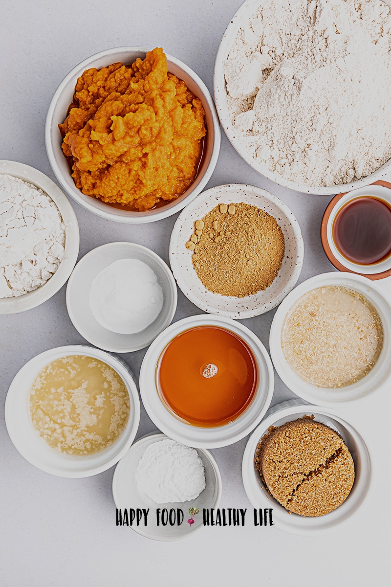 Top view photo of all the ingredients to make vegan pumpkin bread in separate bowls, containing ingredients like pumpkin puree, vegan butter, maple syrup, brown sugar, flax eggs, vanilla extract, oat flour, gluten-free all-purpose flour, pumpkin pie spice, baking soda, and  baking powder.
