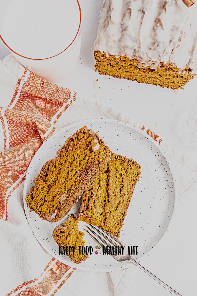 Top view photo of a plate with a slice of vegan pumpkin bread on top. There is a fork, that has cut out a small piece of bread to enjoy. There is a white kitchen towel next to the plate and the remaining pumpkin loaf in the top right of the photo. 
