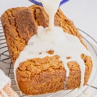 Photo of a loaf of Vegan Pumpkin Bread that is completely cooled on a wire rack. There is a white bowl filled with maple glaze, being poured onto the cooled pumpkin loaf.