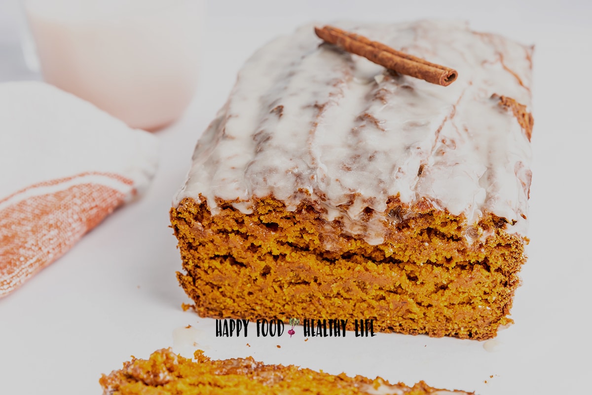 Closeup photo of a vegan pumpkin bread loaf with a maple glaze on top. There is a cinnamon stick on top of the loaf as garnish. The first slice of the loaf has been cut, showing the loaf's moist crumb.