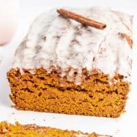 Closeup photo of a vegan pumpkin bread loaf with a maple glaze on top. There is a cinnamon stick on top of the loaf as garnish. The first slice of the loaf has been cut, showing the loaf's moist crumb.