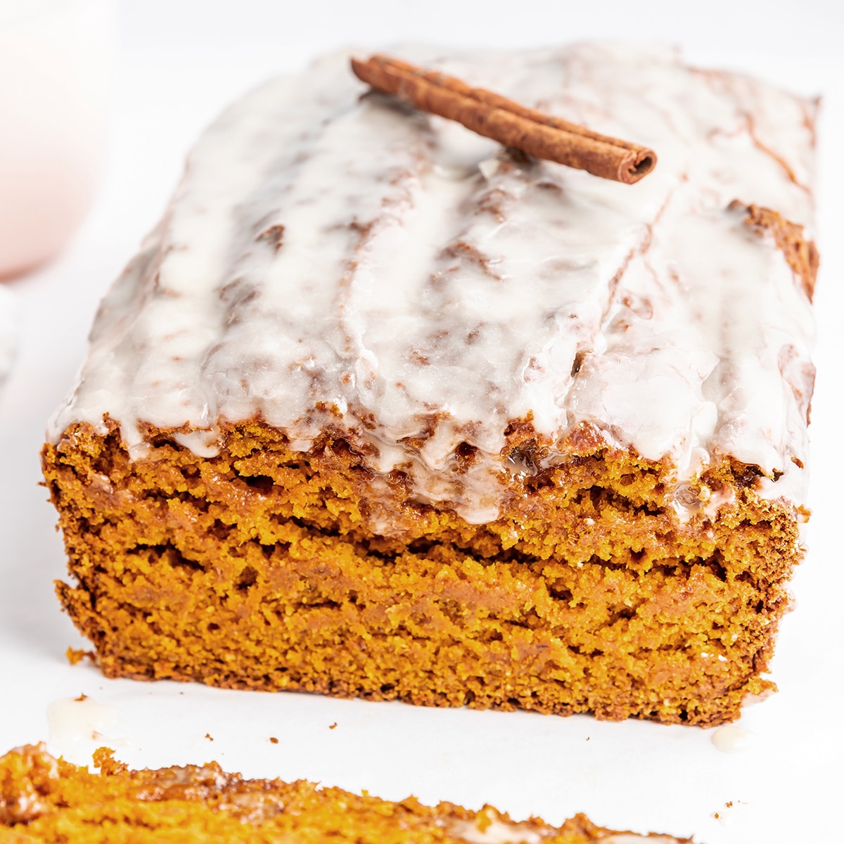 Closeup photo of a vegan pumpkin bread loaf with a maple glaze on top. There is a cinnamon stick on top of the loaf as garnish. The first slice of the loaf has been cut, showing the loaf's moist crumb.