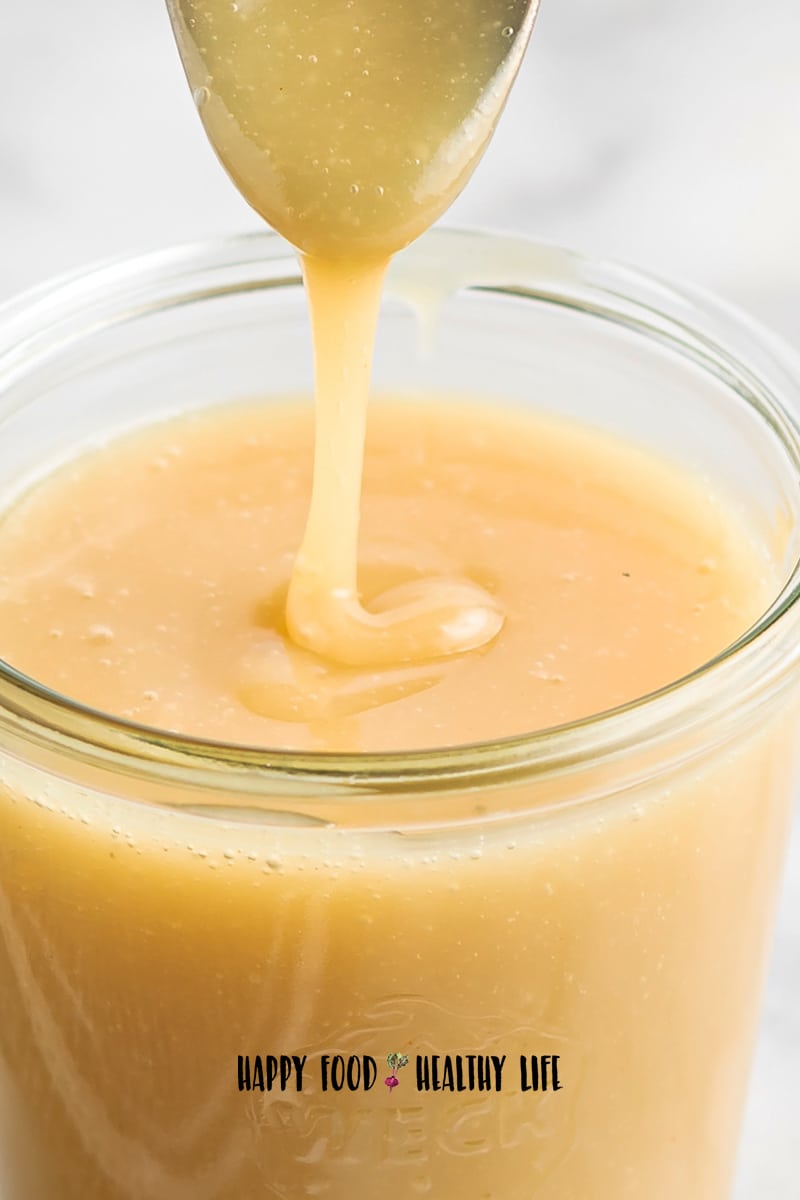 Closeup photo of a spoon that has just been dipped in a glass jar filled with vegan sweetened condensed milk. The spoon is hovering above the glass. It shows how creamy and delicious it looks. 