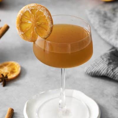 Photo of a coup glass filled with hemlock drink. There is a slice of orange in resting on the edge of the drink. The glass is sitting on a white plate. There are ingredients to make Hemlock drink all around the glass, including star anise and orange slices.