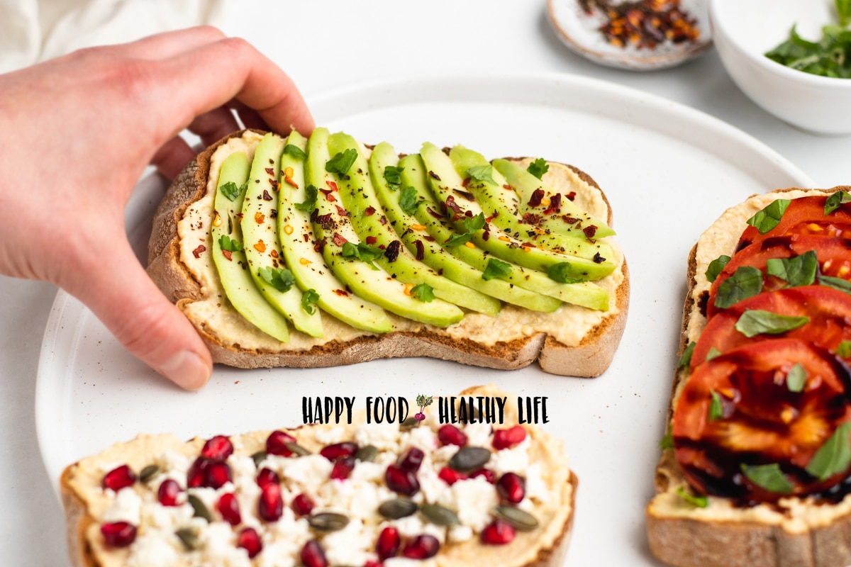 Photo of a hand picking up a piece of hummus toast. This piece has slices of avocados, pepper, red pepper flakes, and cilantro. There are also two other versions of hummus toast by this piece of toast. 