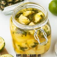 Photo of a glass jar with a glass lid. It is open, and inside it is filled with pickled pineapples. There is a pineapple and a lime in the background, and a wooden cutting board in the foreground.