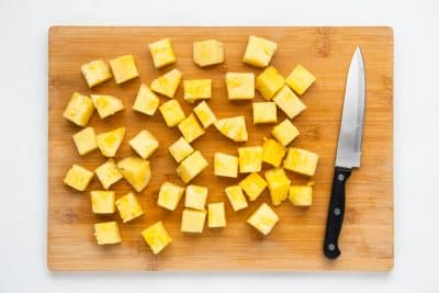 Top view photo of a wooden cutting board with pineapple, chopped into 1-inch chunks on the board. There is a small knife on the cutting board as well.