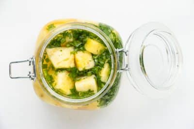 Top view photo of a glass jar filled with pineapple chunks, chopped cilantro, and diced jalapeno in the jar, and filled to the top with pickling liquid. It is ready to chill until ready to enjoy.
