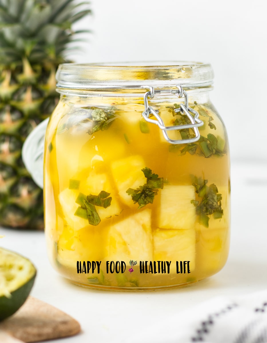 Photo of a glass jar with a glass lid. It is closed, and is filled with pickled pineapple. In the background is a pineapple and in the foreground is a cutting board with half a lime. 