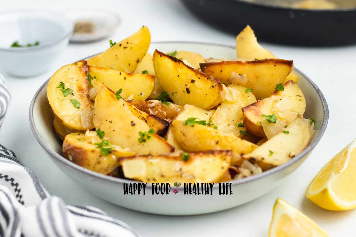 Photo of vesuvio potatoes in a large white bowl, sprinkled with freshly chopped parsley. There are lemon wedges and a kitchen towel in the foreground and small white bowls in the background.