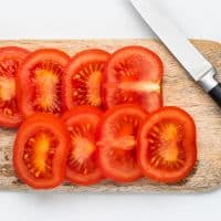 Top view photo of a wooden cutting board, a small knife, and slices of a red, juicy tomato.